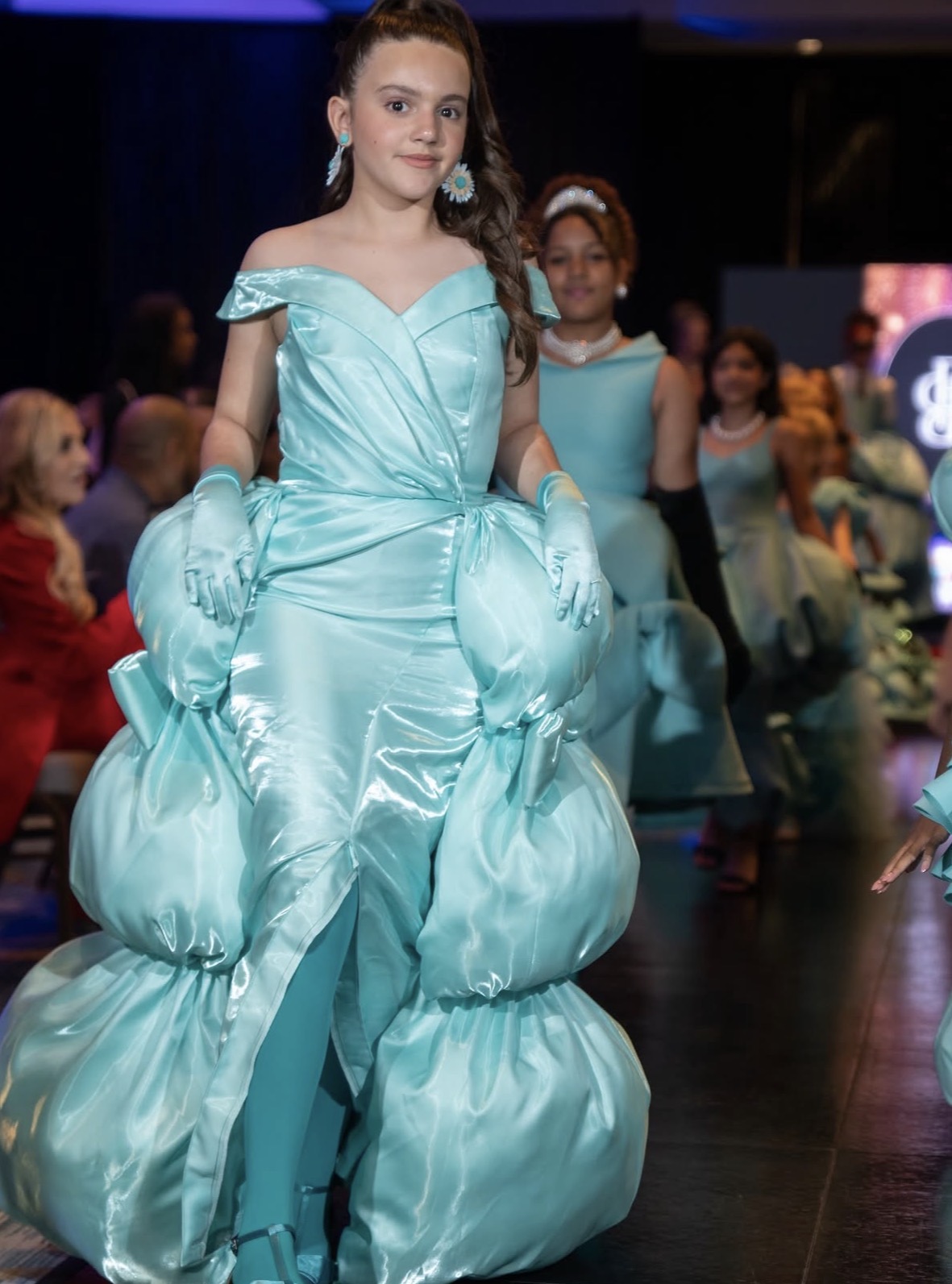 Girl in a light blue dress with puffy skirt and gloves, walking on stage with other children in background.