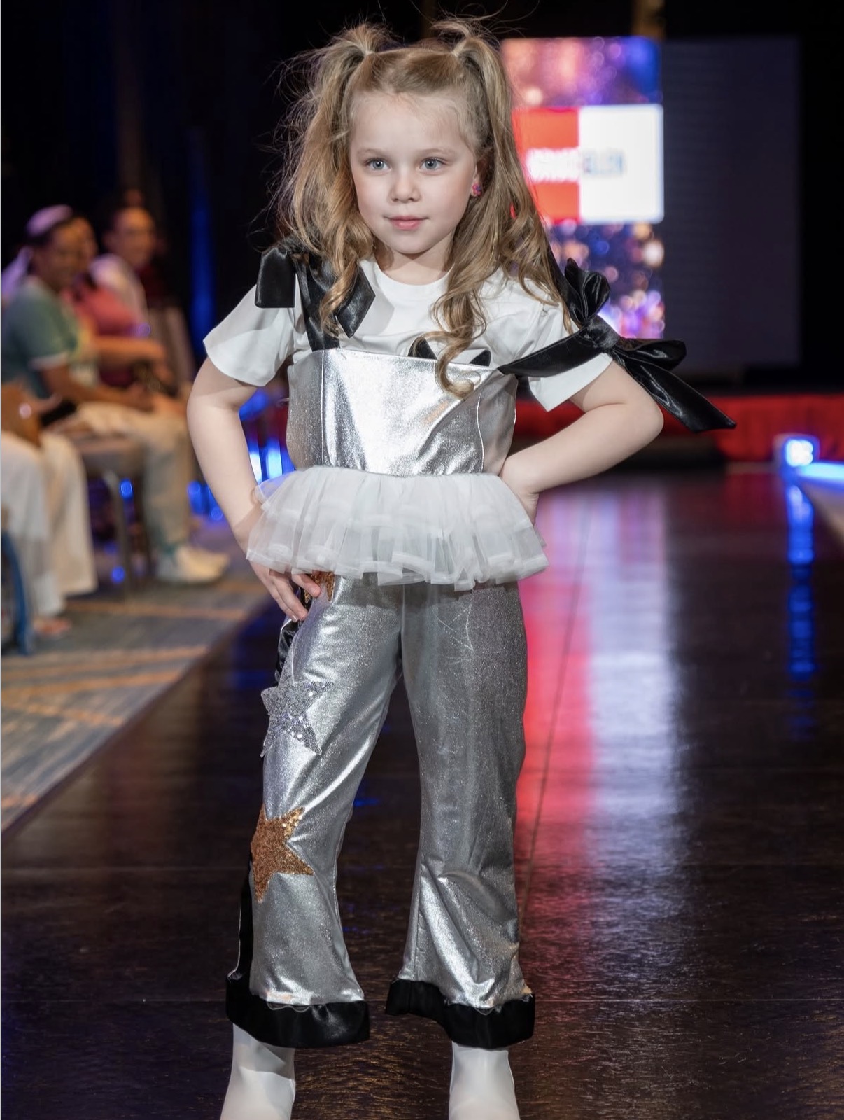 Young girl with long hair in a silver and black outfit walking on a runway, audience in background.