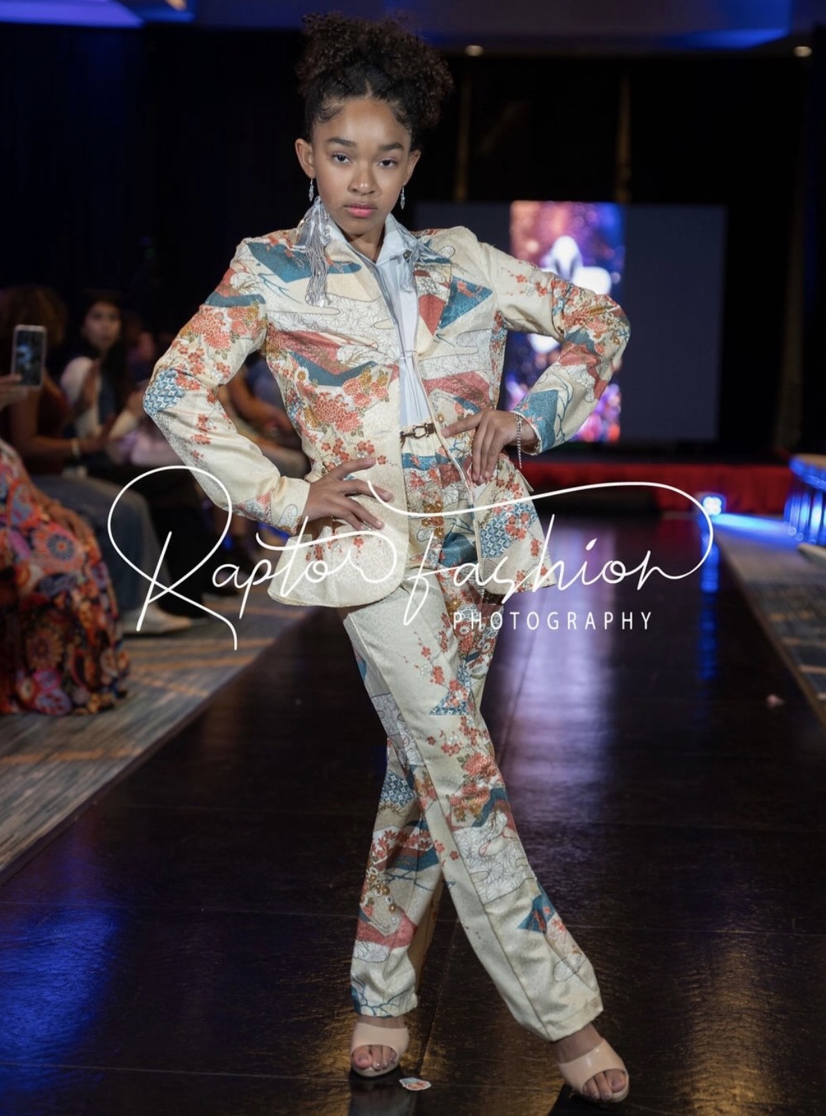 Young girl walking on runway in colorful patterned outfit with hands on hips, dark background, audience in distance.