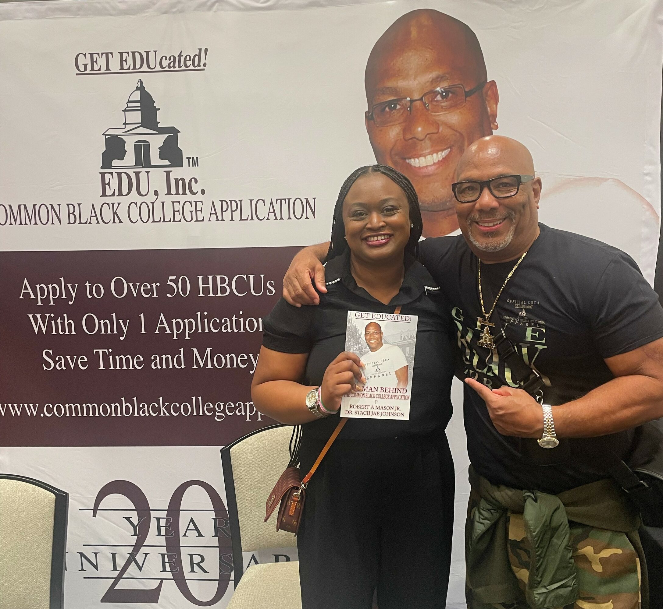 Two people smiling, one holding a brochure, standing in front of a banner with a large man's face and text about college application.