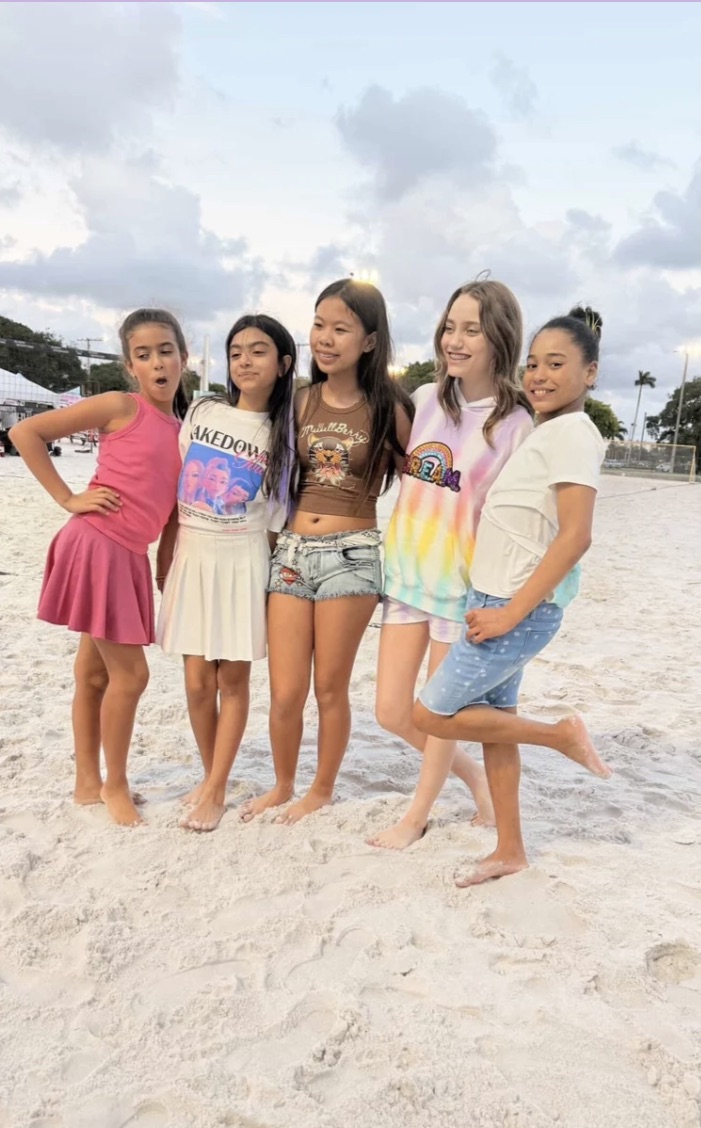 Five girls standing on a sandy beach under a cloudy sky, posing for a photo, some with hands on hips or crossed arms.