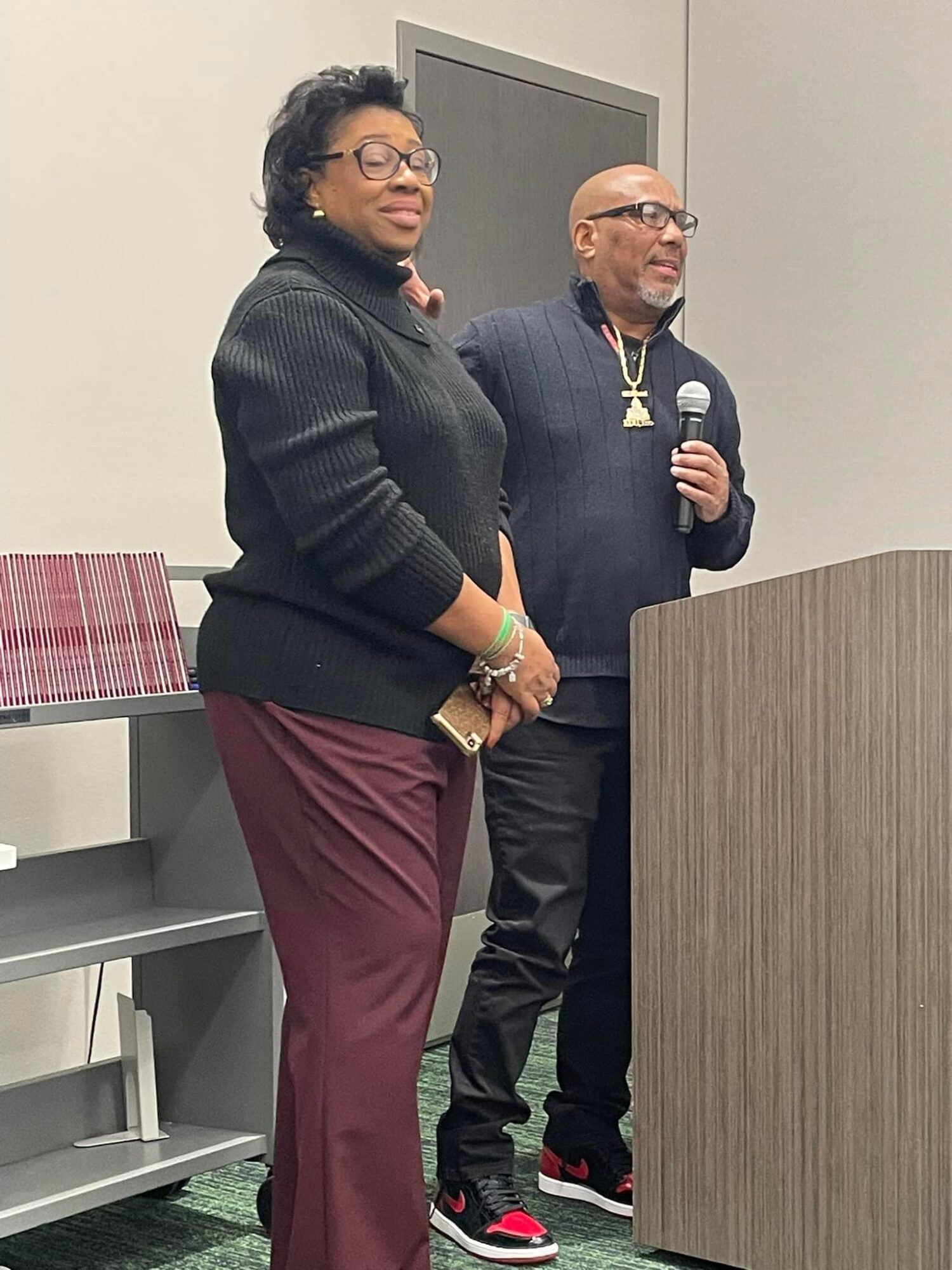 Two people standing behind a podium, one holding a microphone, in a room with a gray wall and furniture.