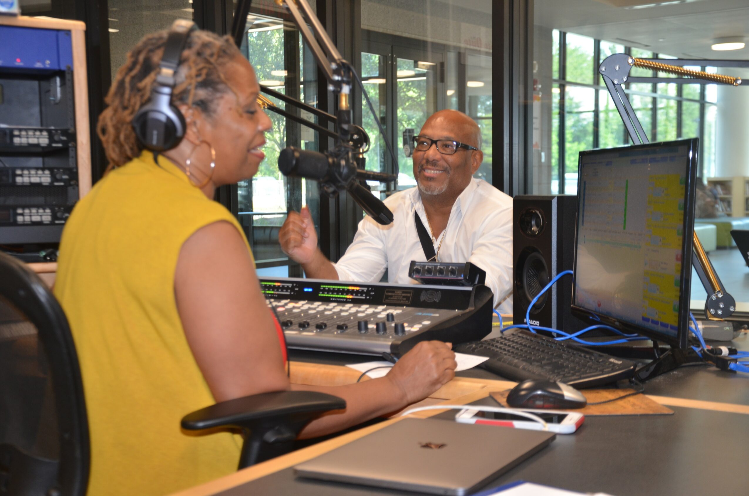 Two people in a radio studio, one speaking into a microphone, the other smiling and gesturing, with audio equipment and a computer.