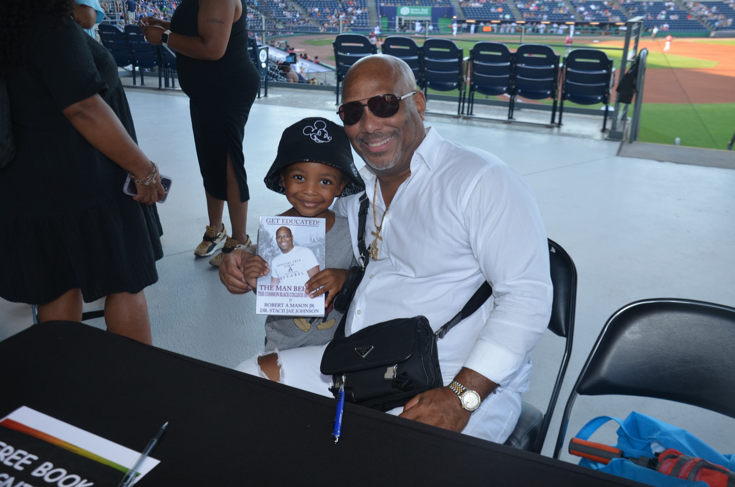 Man and child sitting at a table, smiling, with a baseball field in the background, holding a booklet.