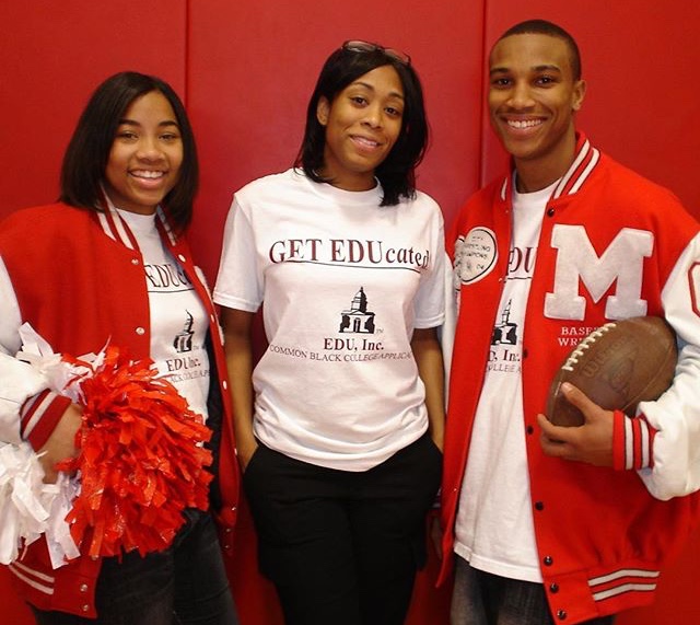 Three people standing against a red background, two wearing red jackets and one in a white shirt, smiling.
