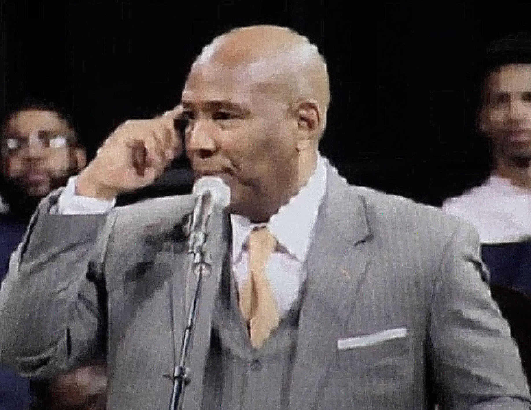 Man in a gray suit speaking into a microphone, with people in the background, in a formal setting.