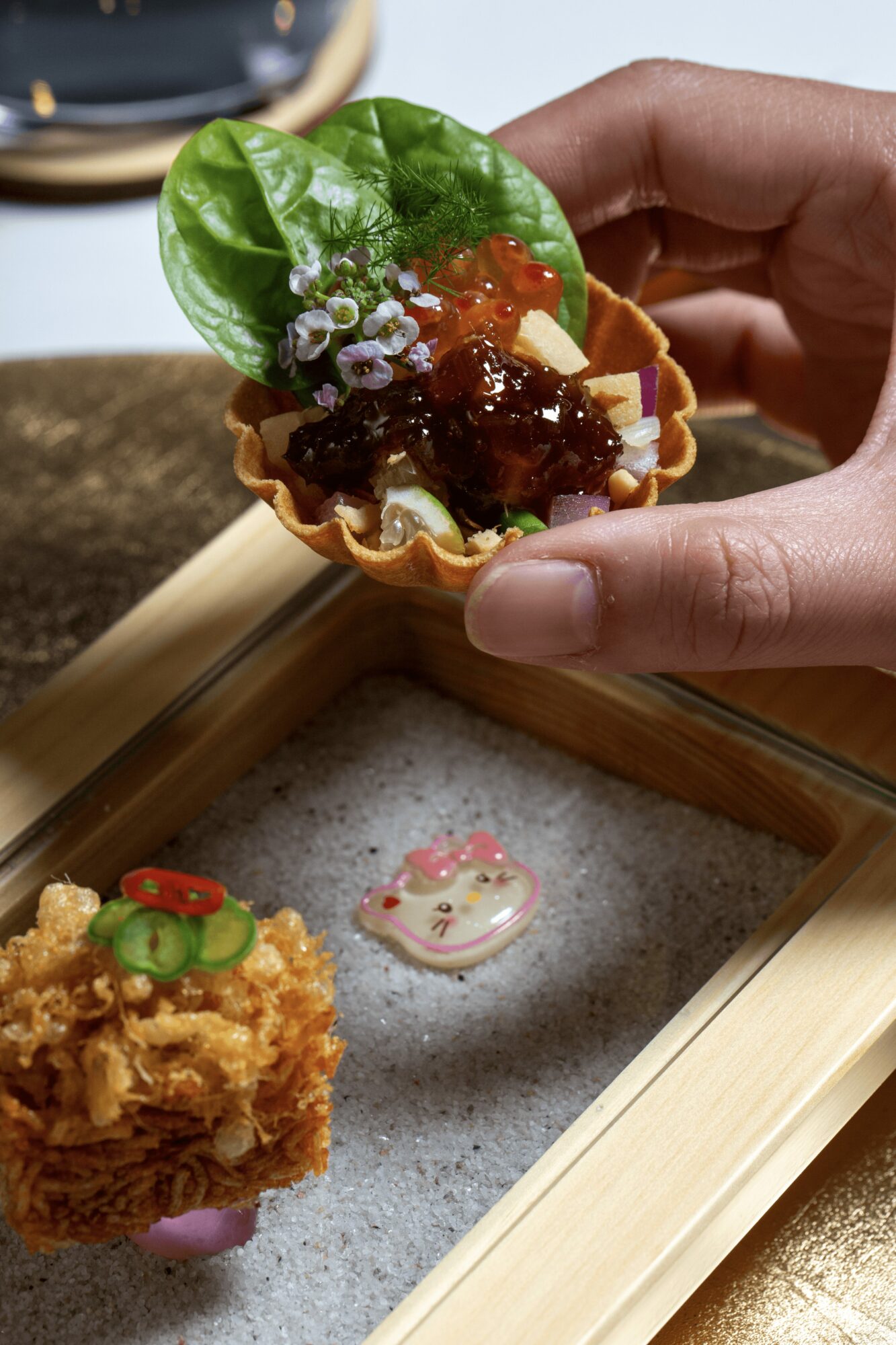 Hand holding a small edible cup with lettuce, sauce, and toppings, above a wooden tray with a decorated cookie and fried food.