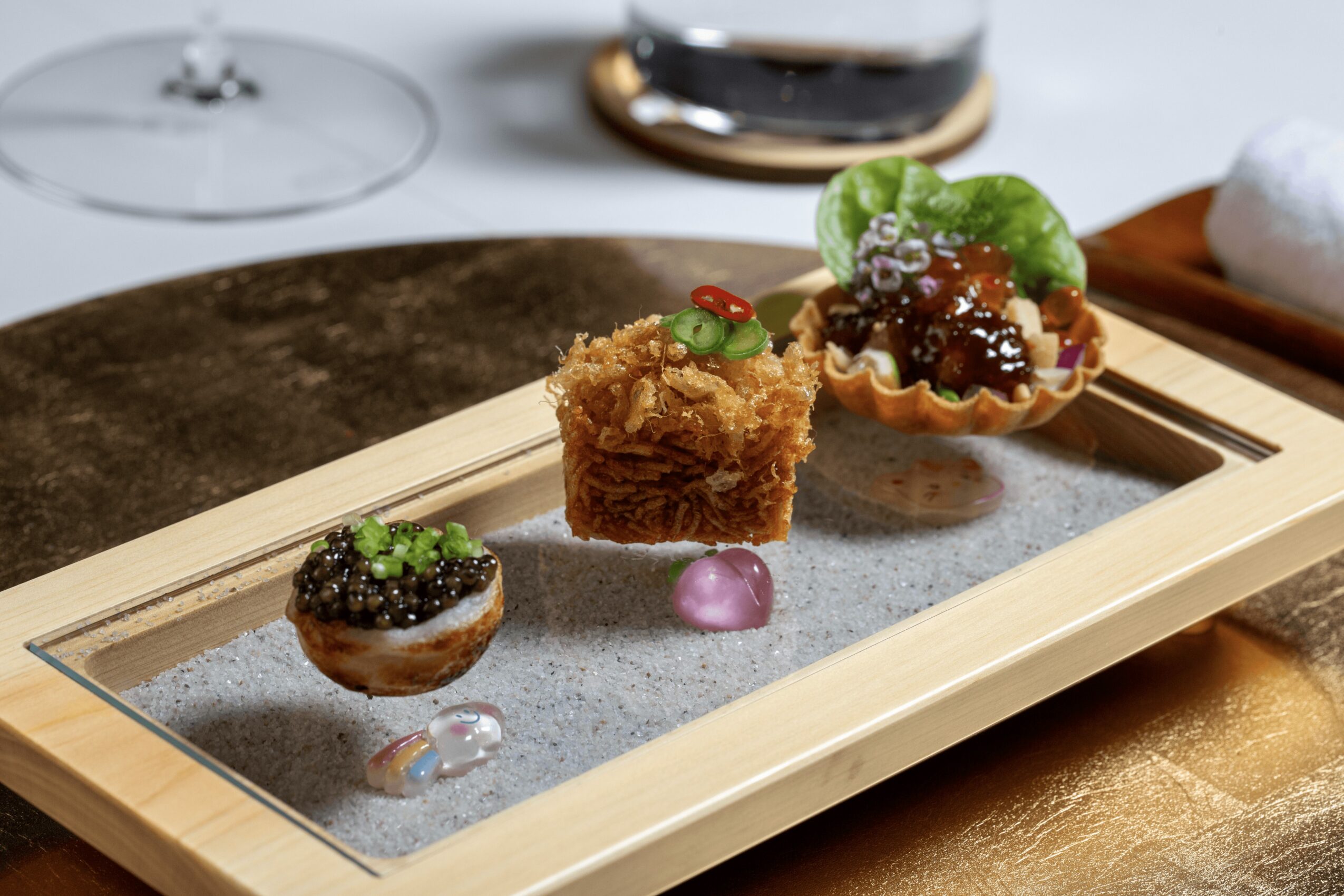 Three small desserts on a wooden tray with a glass of dark beverage in background, on a white tablecloth.