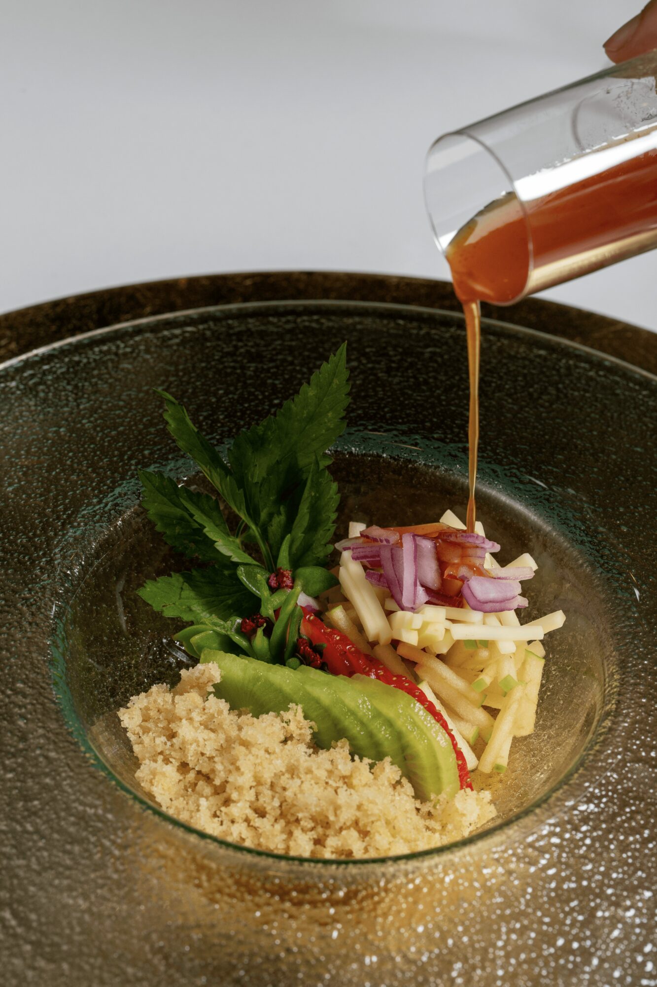 Pouring sauce over a plated dish with vegetables and grains in a glass bowl.