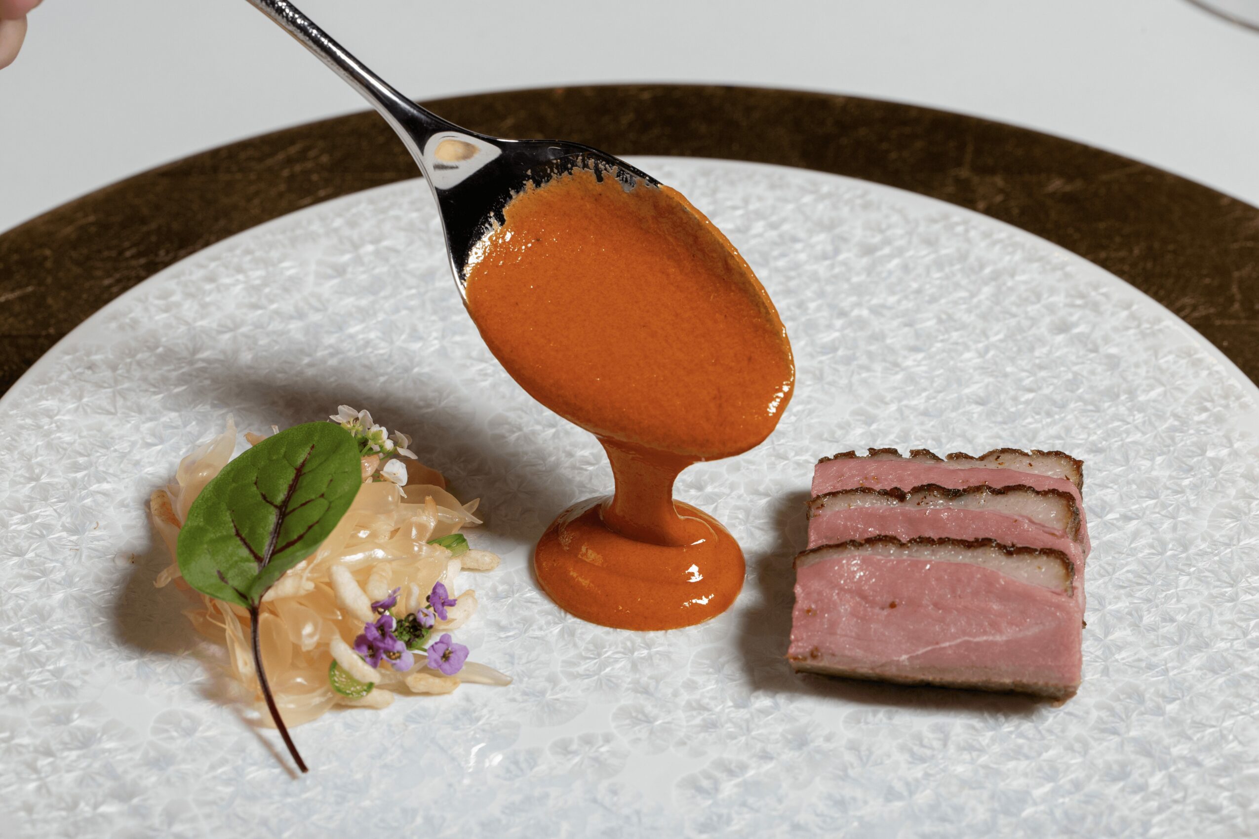 Spoon with orange sauce above a plate with a small green leaf, edible flowers, and a rectangular piece of meat.