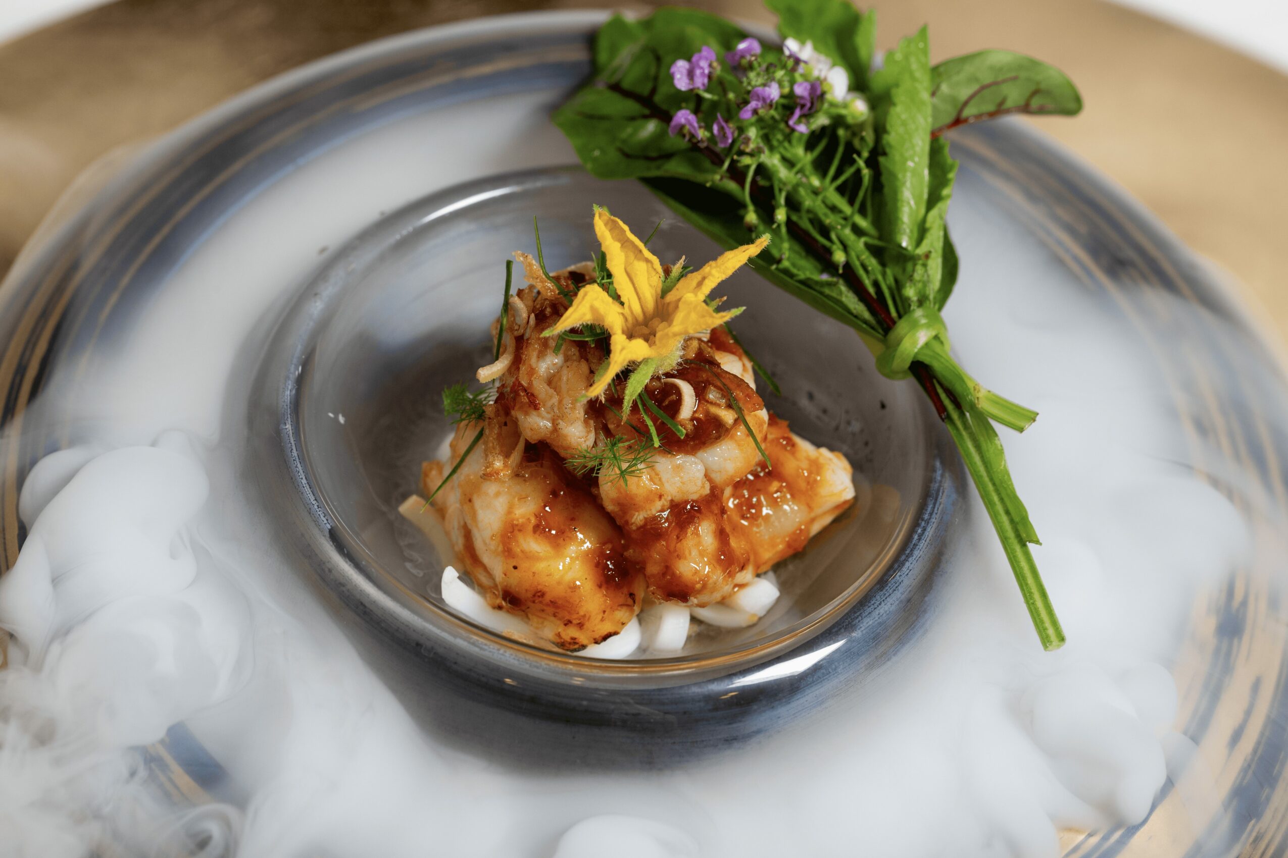 A plated dish with a piece of cooked fish topped with herbs, garnished with a sprig of green leaves and purple flowers, on a glass plate with fog effects.