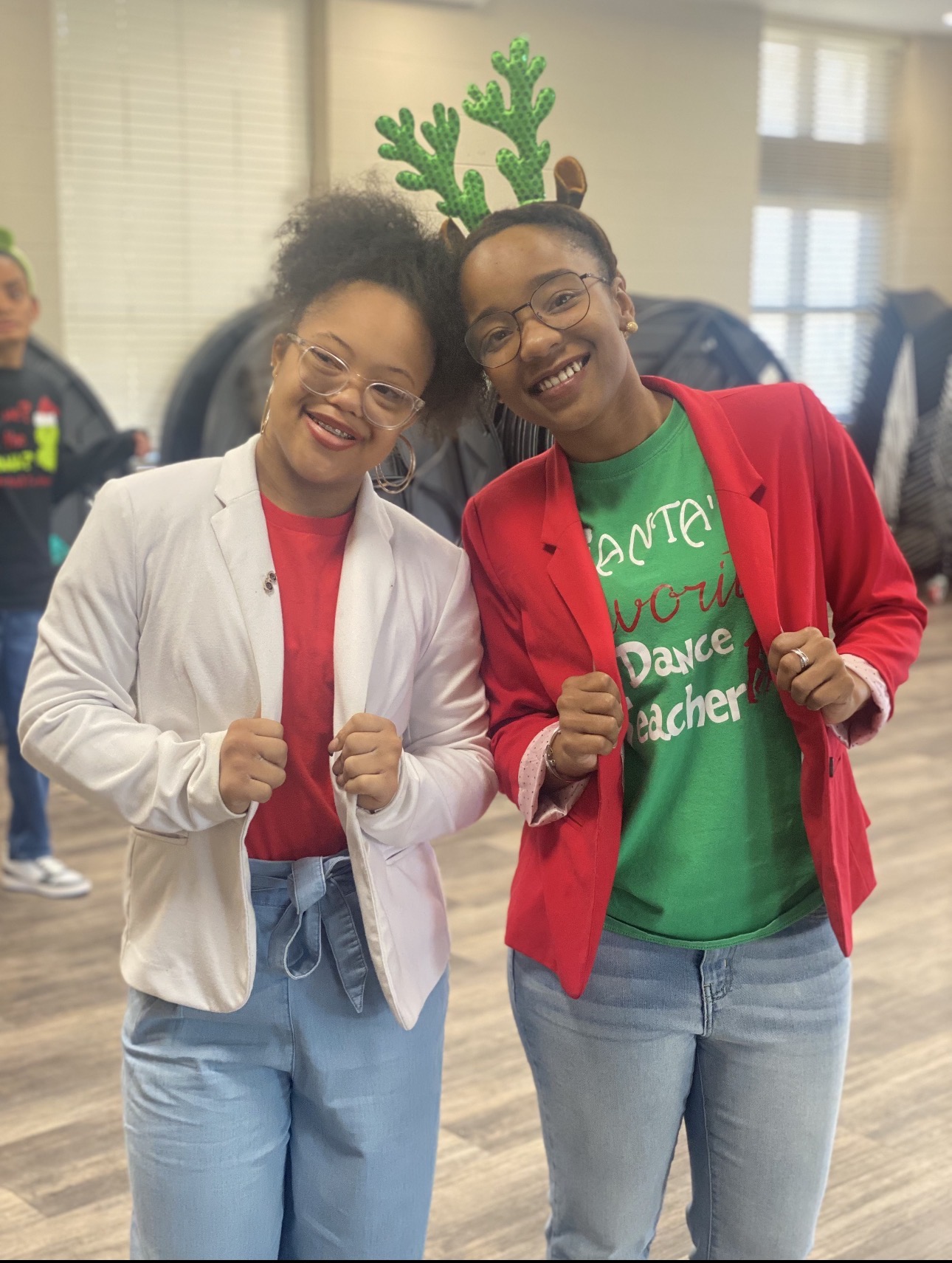 Two women smiling, one wearing reindeer antlers, in a room with other people, celebrating together.