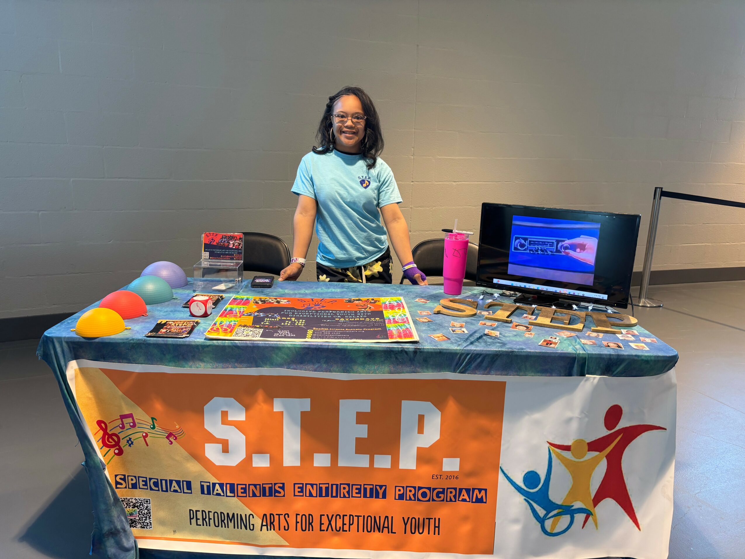 Girl standing behind a table with a banner reading S.T.E.P. performing arts program, displaying games and a laptop.
