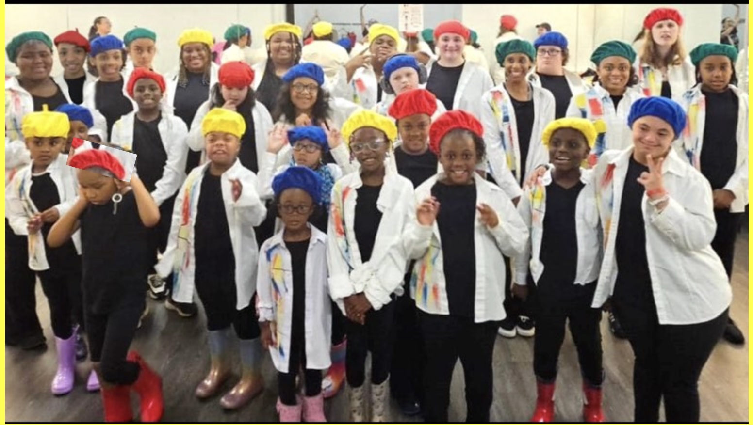 Group of children wearing colorful hats and white coats, standing together indoors, smiling at the camera.