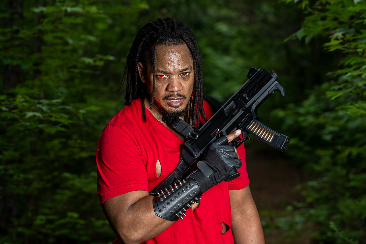 Man with dreadlocks in a red shirt holding a black firearm in a forest setting.
