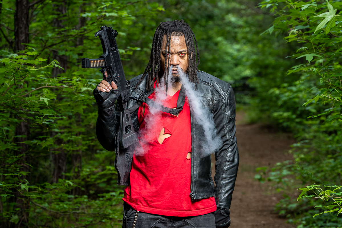 Man with dreadlocks in a forest, wearing a black leather jacket and red shirt, holding a gun, with smoke around his face.