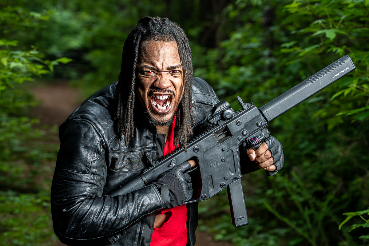Person with dreadlocks and leather jacket holding a large gun outdoors, shouting or yelling, surrounded by green foliage.