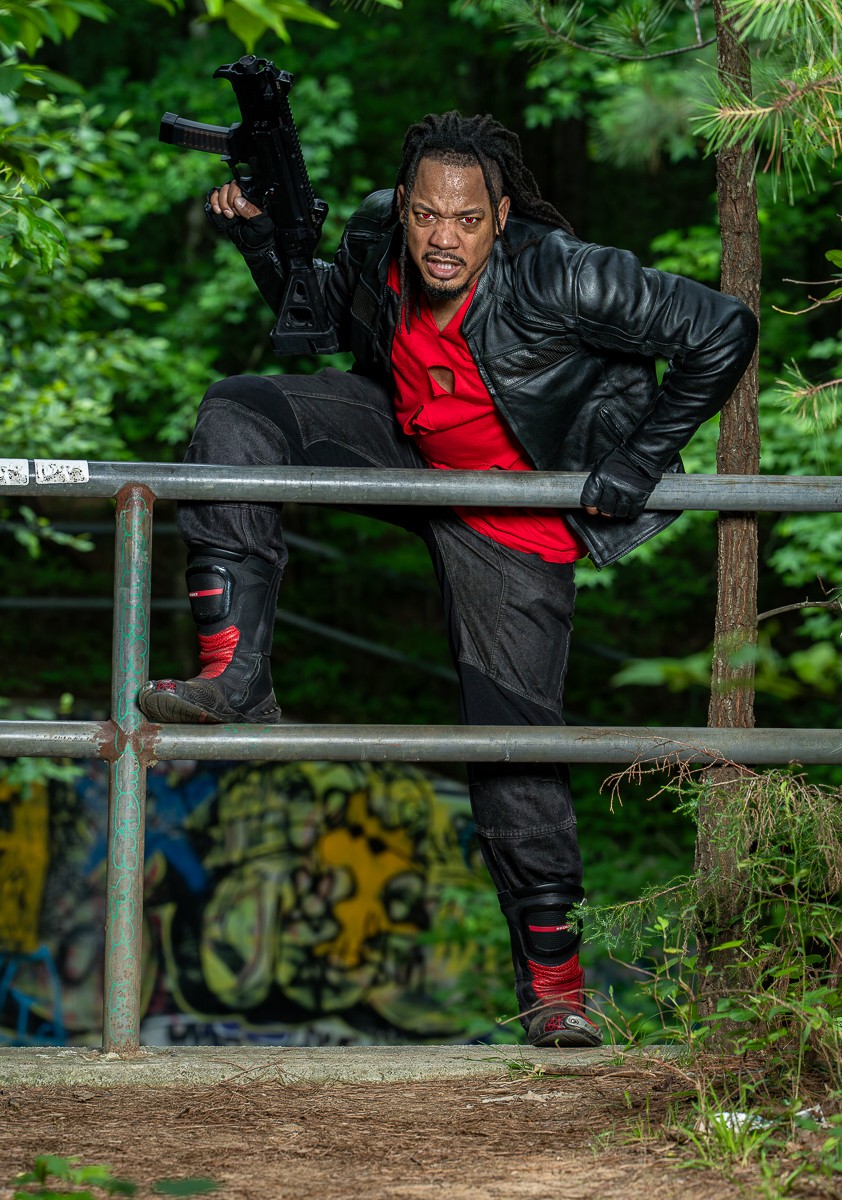 Man with dreadlocks in a black leather jacket and red shirt poses outdoors with a gun, surrounded by green trees.