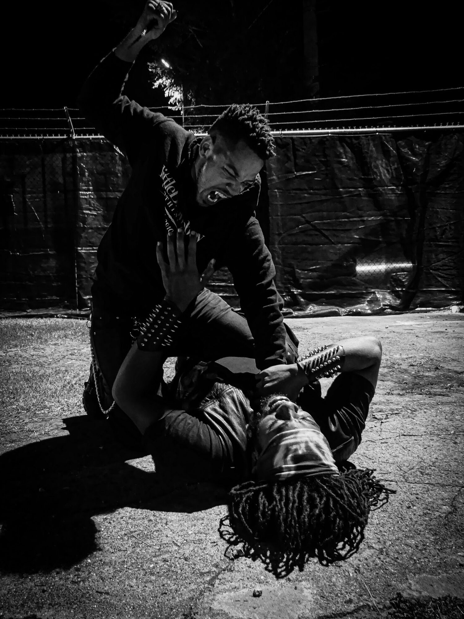 Two people fighting on the ground outdoors at night, one standing and one lying down, with a fence in the background.