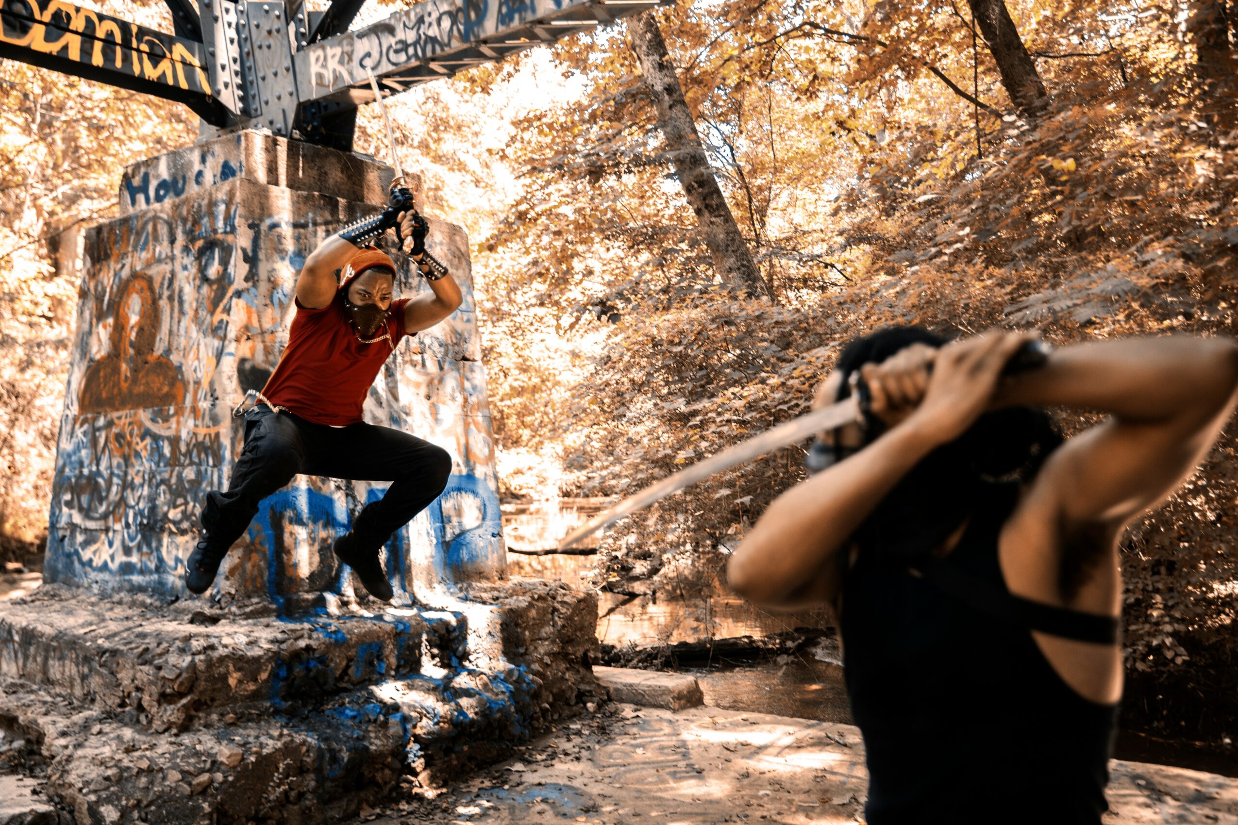 Two people outdoors near graffiti-covered concrete stairs, one sitting and taking a photo, the other holding a stick or pole, surrounded by trees with autumn leaves.