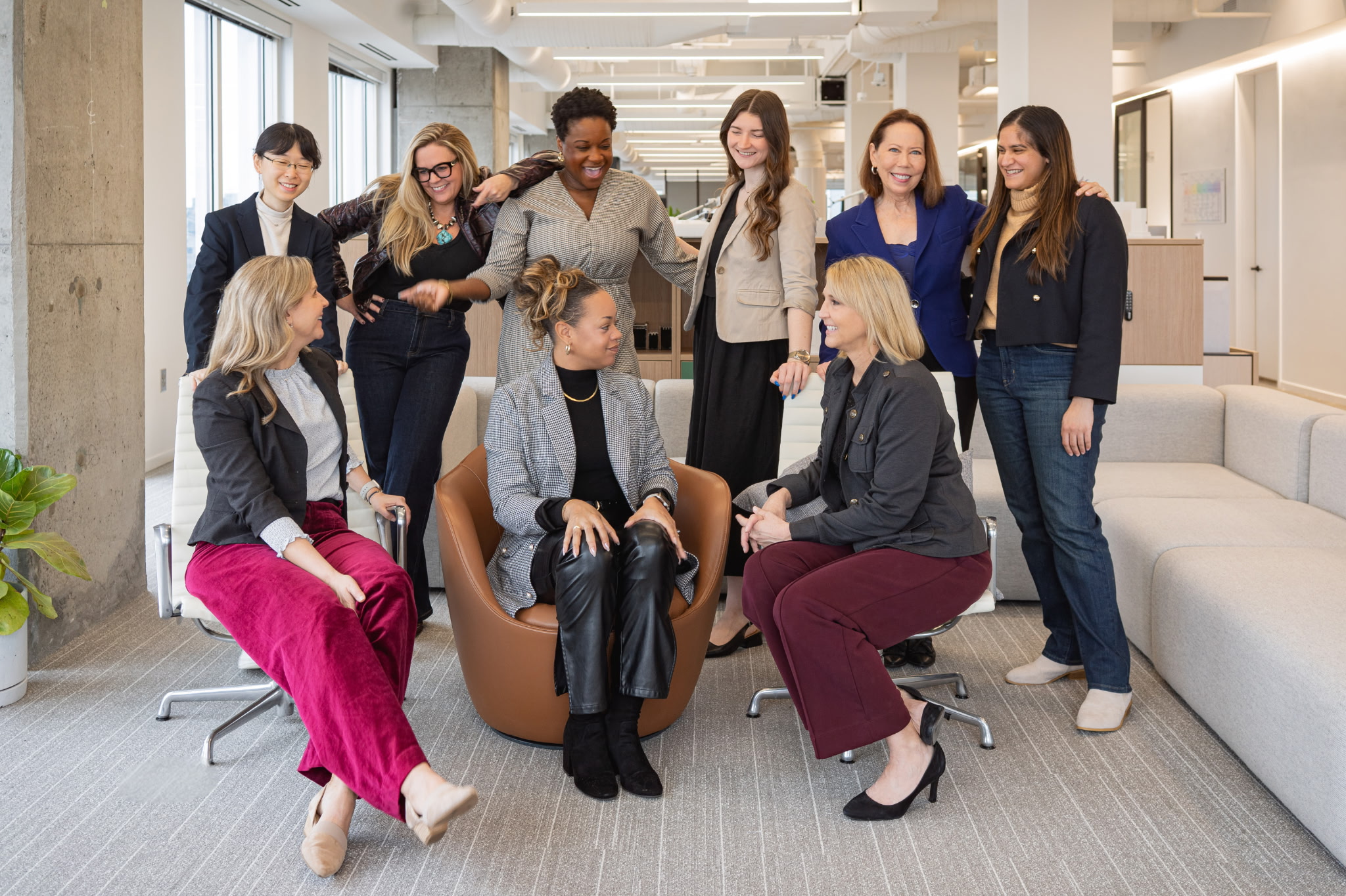 Group of nine diverse women in a modern office, some sitting and some standing, engaging in conversation and smiling.