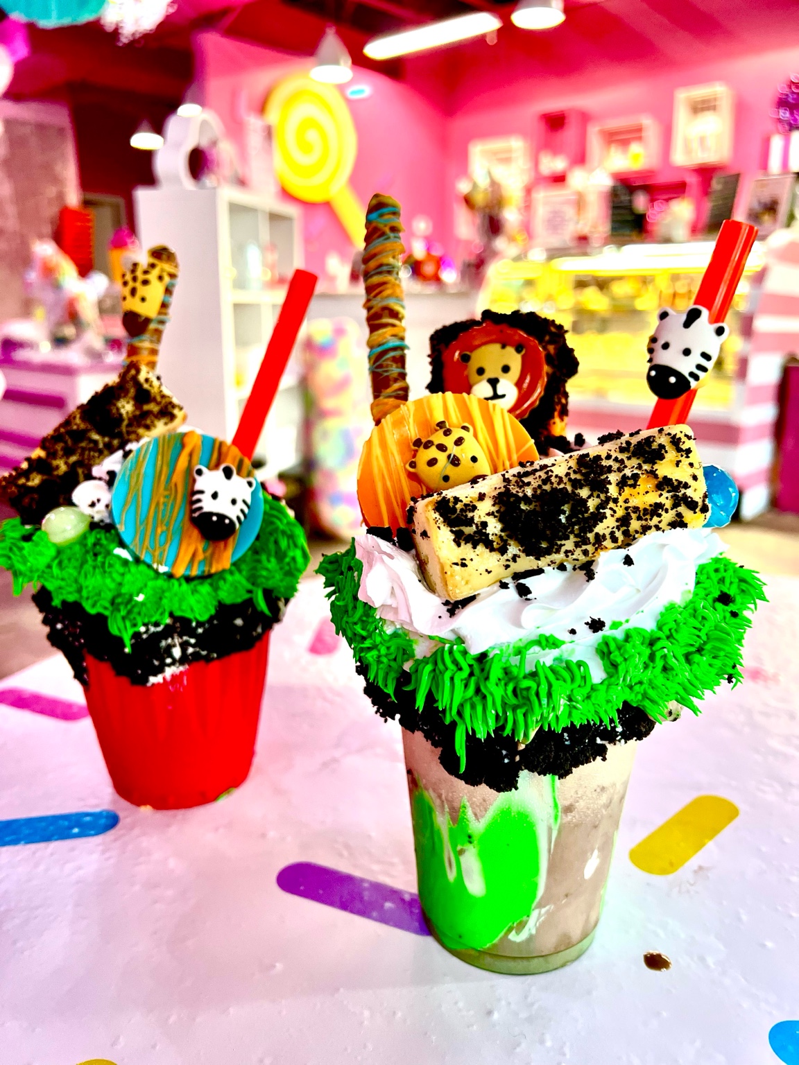 Two colorful milkshake desserts with toppings, candies, and cookies in a decorated store, pink walls, and shelves in background.