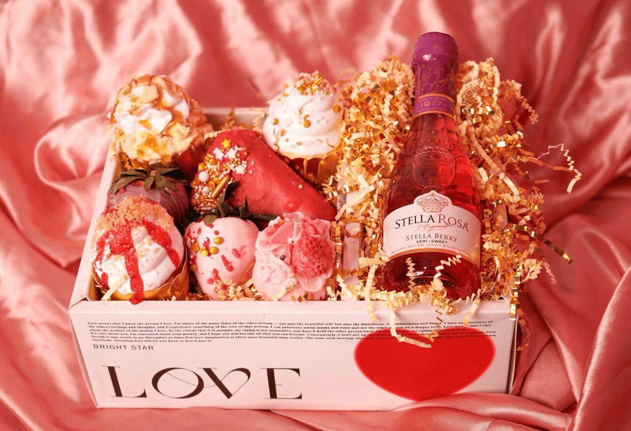 Box of assorted colorful desserts and a small bottle of pink sparkling wine on pink satin fabric.