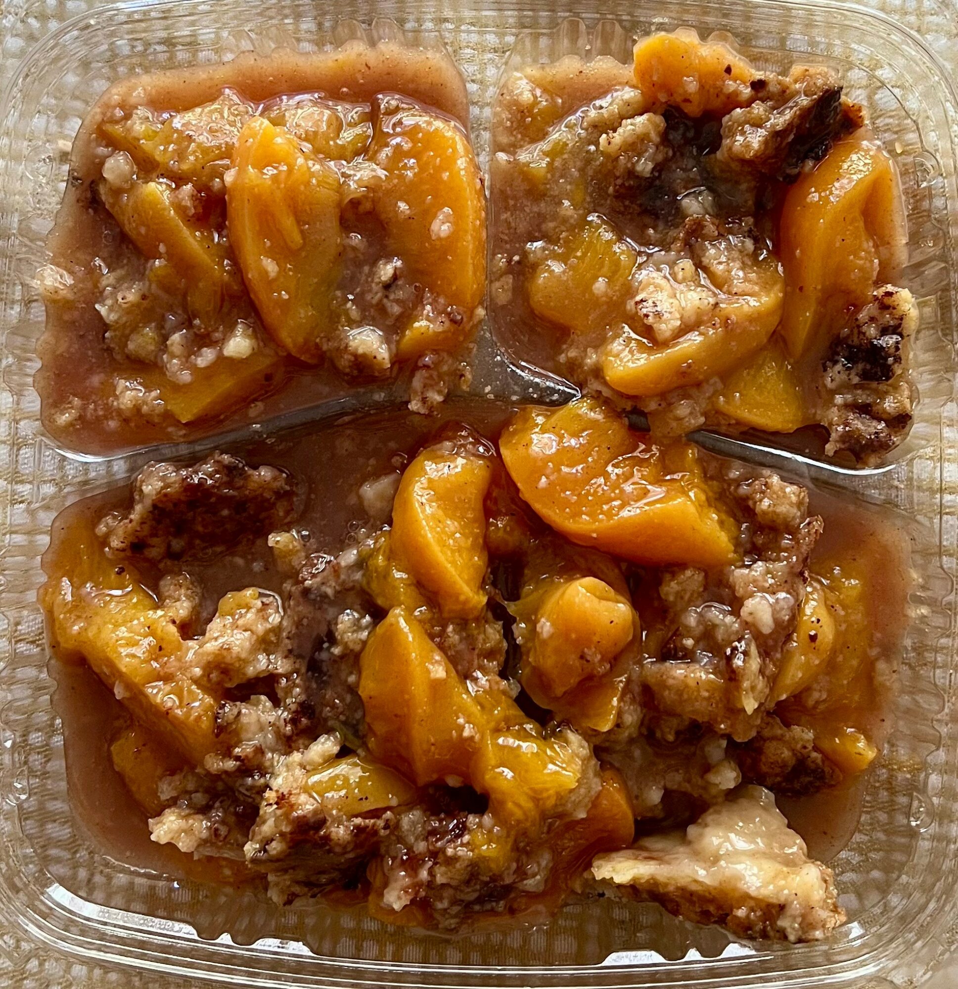 Four pieces of peach cobbler in a clear container with a spoon, showing golden peaches and crumbly topping.