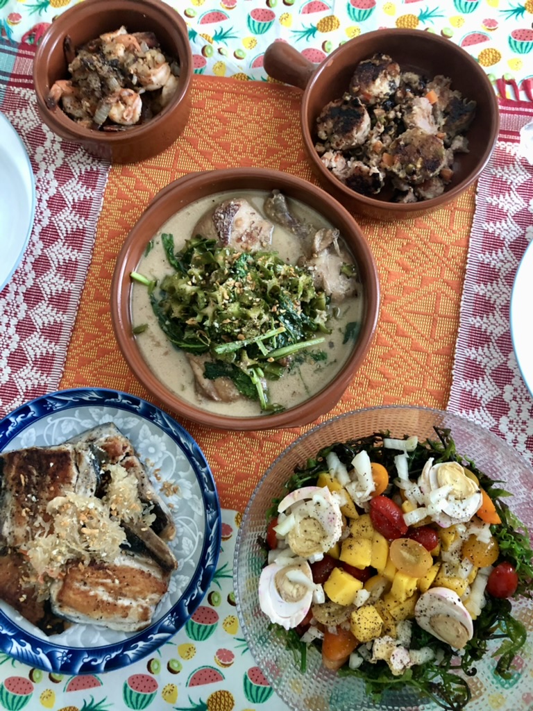 Assorted dishes on a colorful tablecloth, including a green vegetable dish, a salad with bananas and cherry tomatoes, and baked items.
