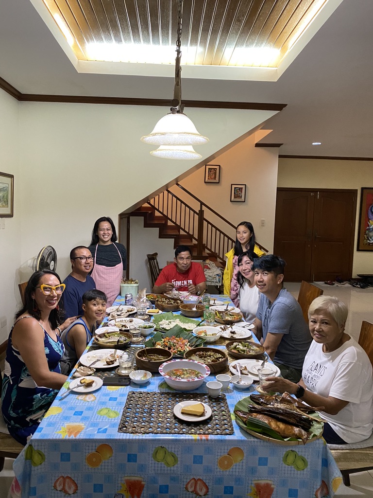 Group of nine people gathered around a dining table with food, in a room with a staircase and ceiling light.