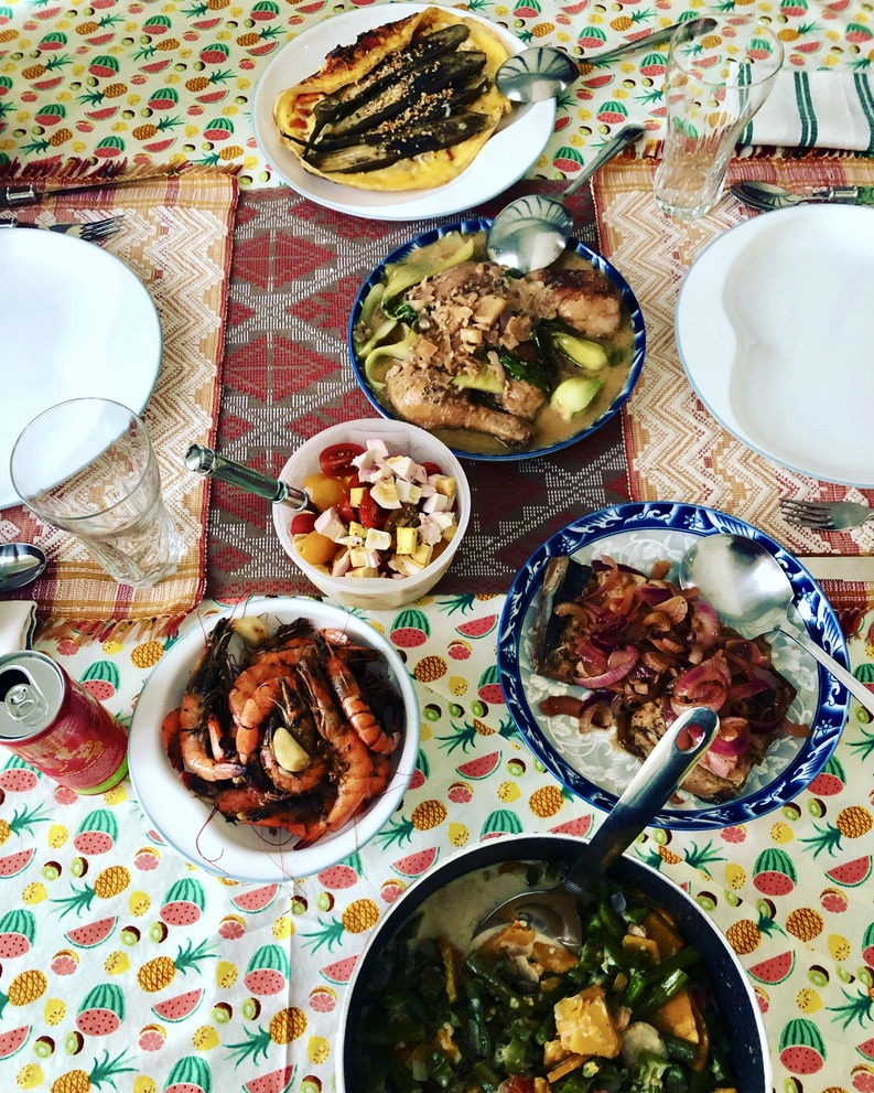 Table with various dishes including grilled meat, vegetables, and salad, set on a colorful patterned tablecloth with empty white plates and glasses.