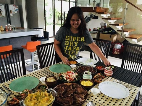 Woman smiling at a table with various dishes, in a modern kitchen with stairs in the background.