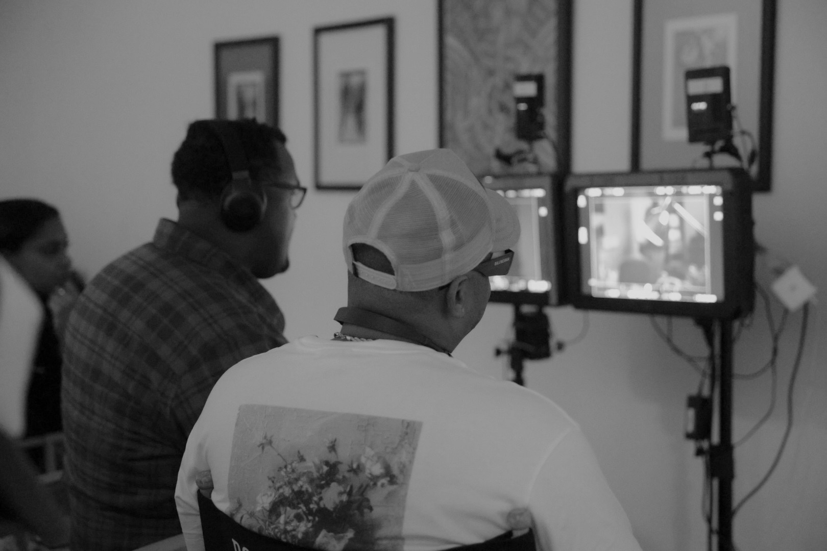 Two people are filming or photographing in a gallery with framed pictures on the wall, one wearing headphones and a cap.