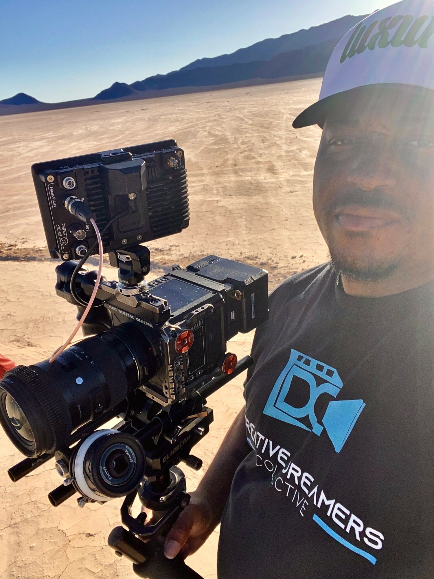 Person wearing a cap and T-shirt with camera equipment in a desert landscape with mountains in the background.