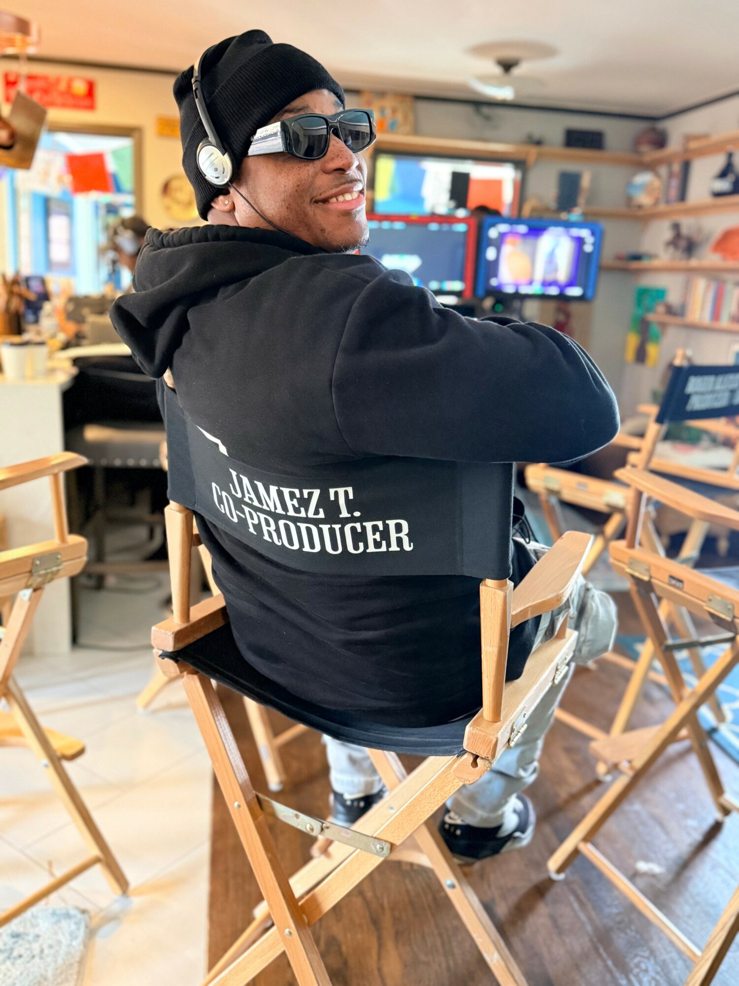 Person wearing headphones and sunglasses, sitting in a studio or workspace with computers and shelves, smiling and looking to the side.