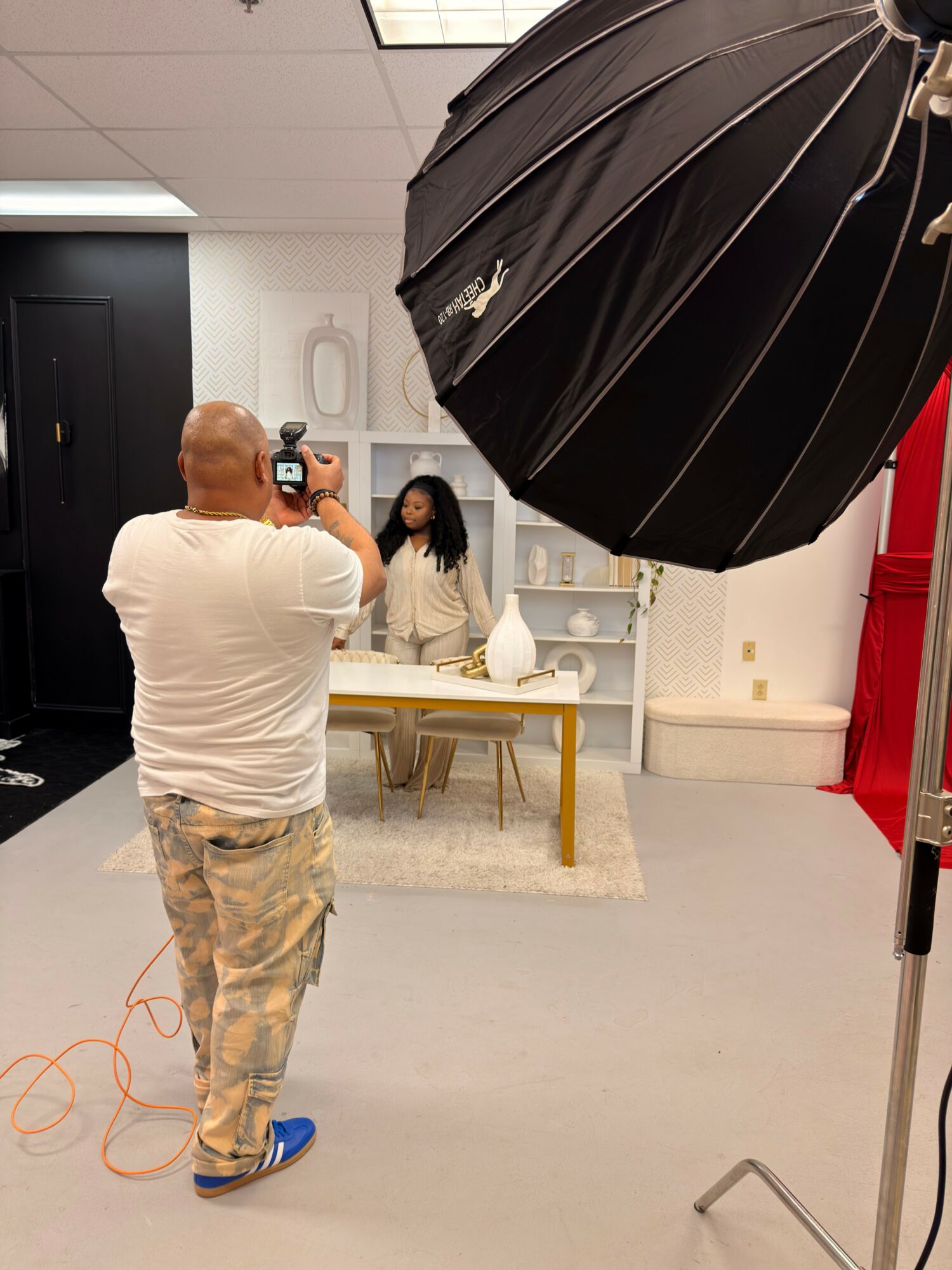 Person taking a photo in a room with a large black umbrella, white walls, and shelves with various items.