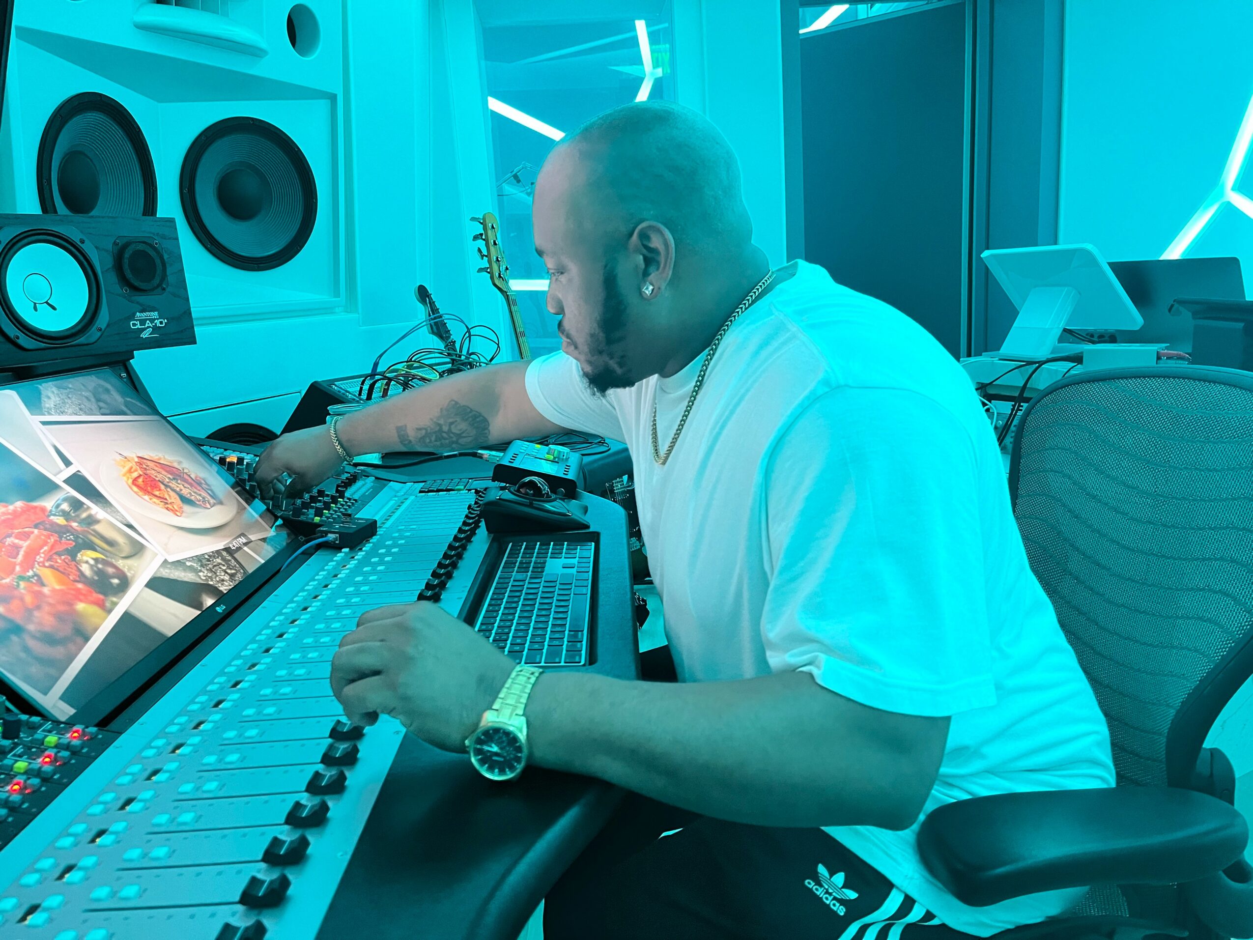 Man working at a music production console with speakers and computer screens in a modern studio, illuminated by blue lighting.