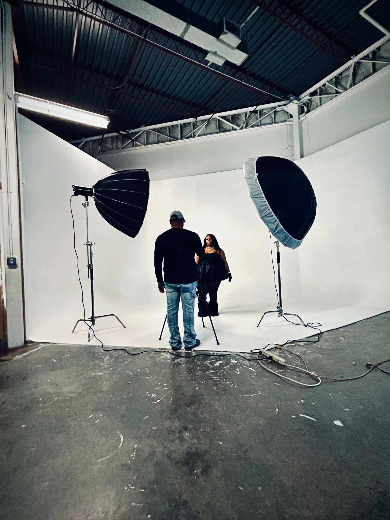 Two people stand in a photography studio with large softbox lights, white curved backdrop, and industrial ceiling.