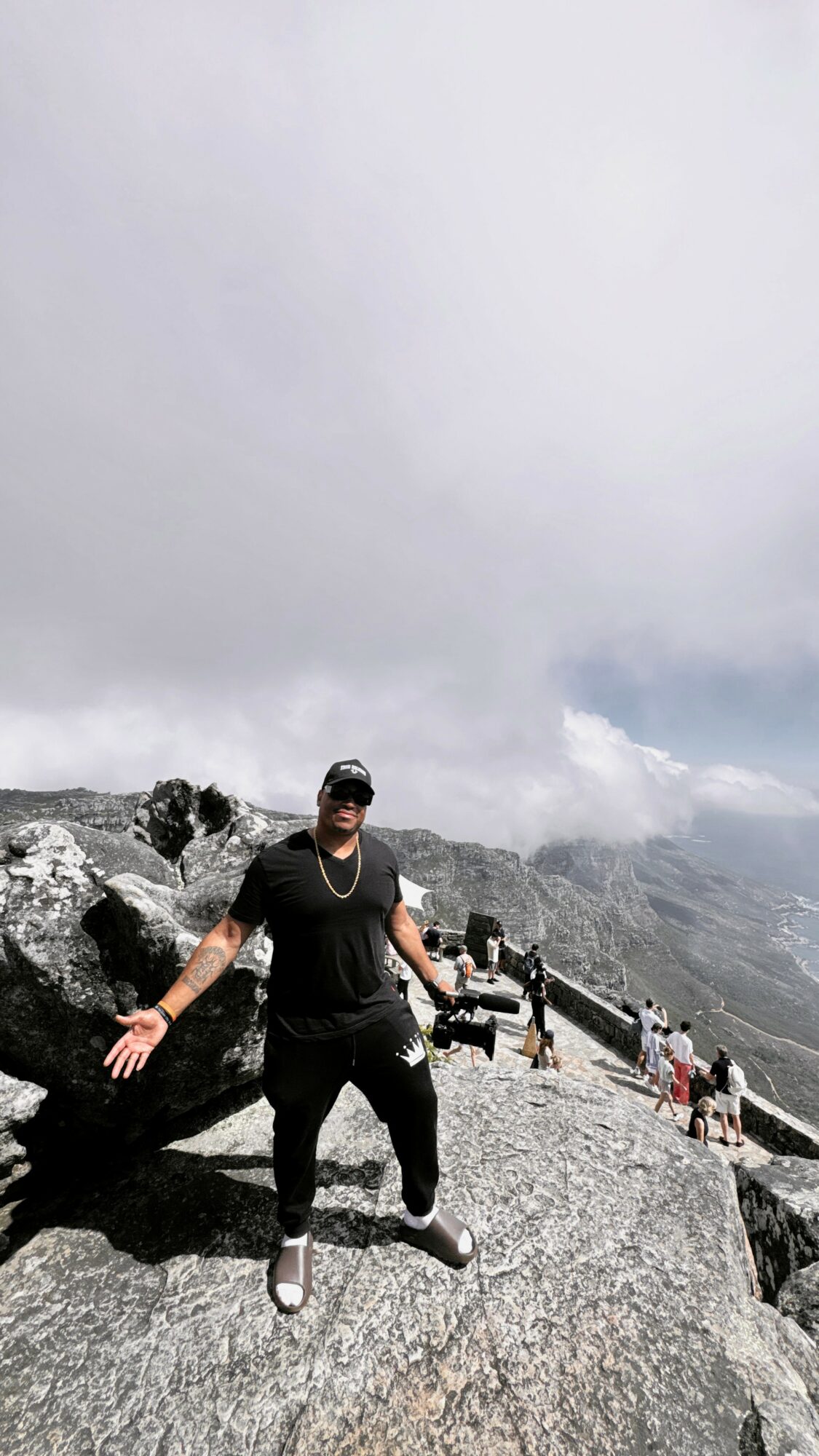Person standing on rocky mountain with cloudy sky and distant mountain views, wearing dark clothing and sunglasses.