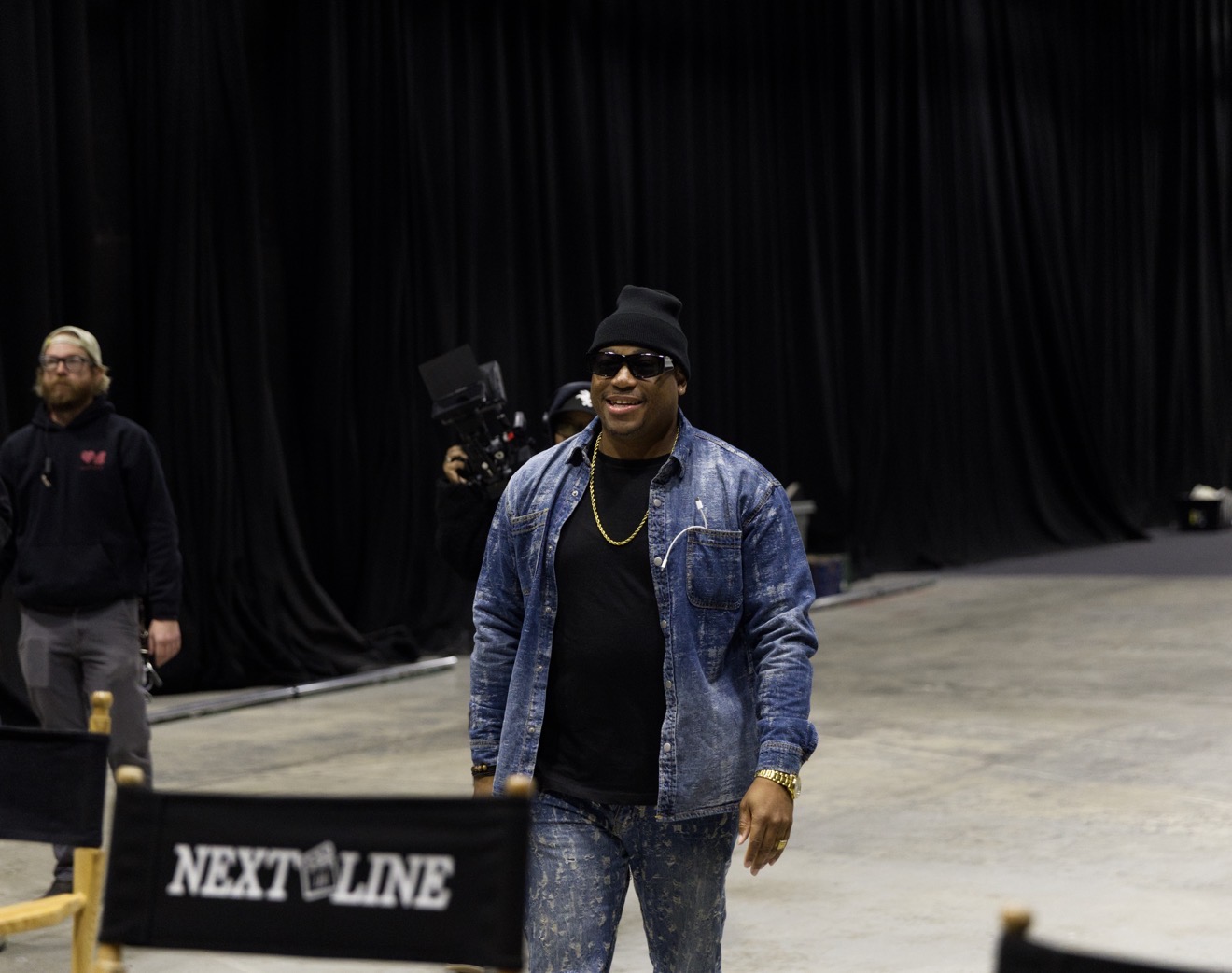 Person wearing sunglasses, beanie, denim jacket, and black shirt walking in an indoor space with black curtains.