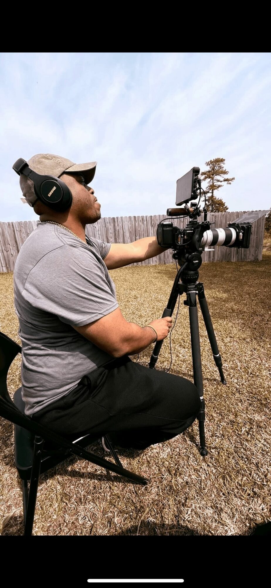 Man with headphones operating camera on tripod outdoors, cloudy sky background.
