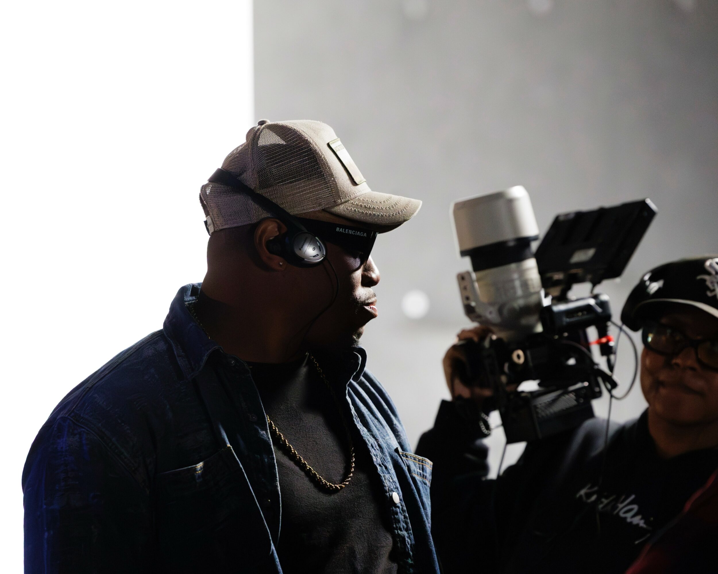 Person wearing a cap and headphones, with a camera operator nearby, in a dimly lit setting.