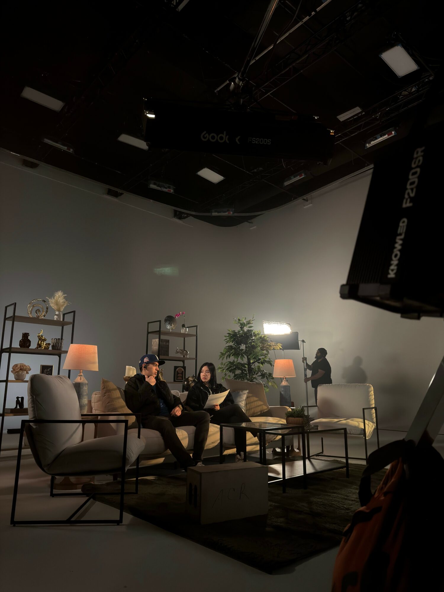 Two people sit on a sofa in a studio with lamps, plants, and a large white backdrop, preparing for a recording or interview.