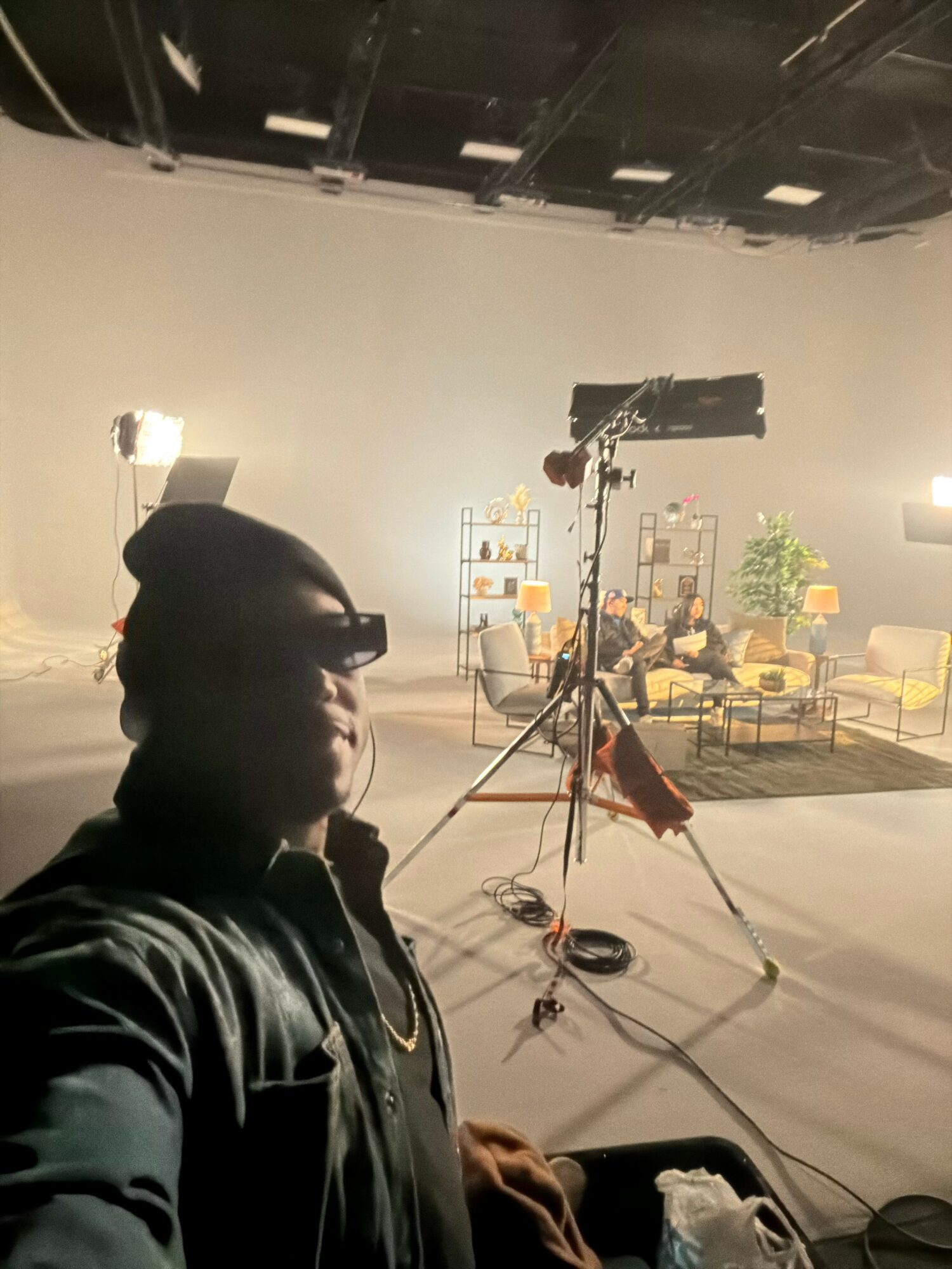 Person taking selfie in a studio with set furniture, lighting, and camera equipment in the background.