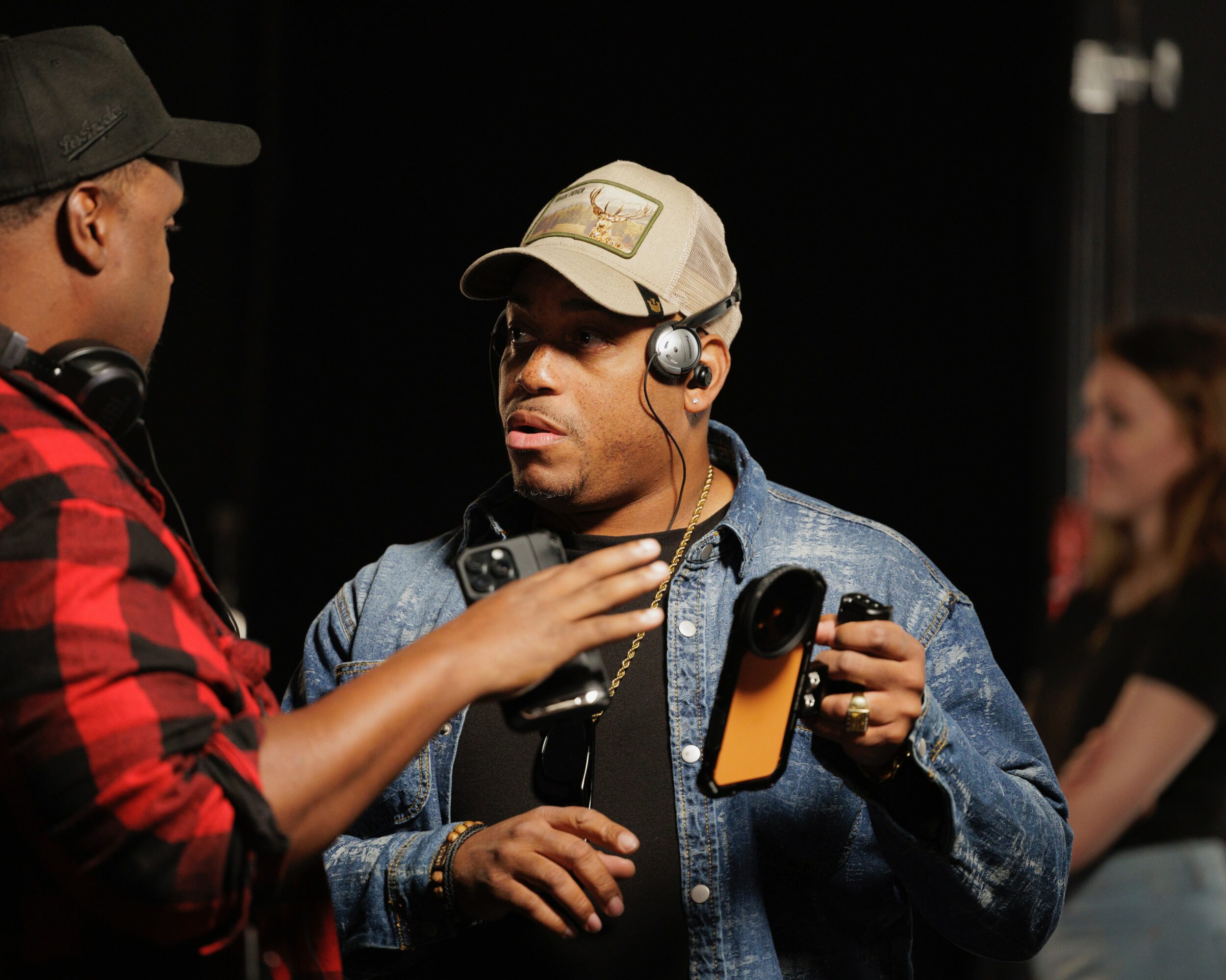 Two men in conversation, one wearing a cap and headphones, the other in a red and black checkered shirt, in a dark setting.