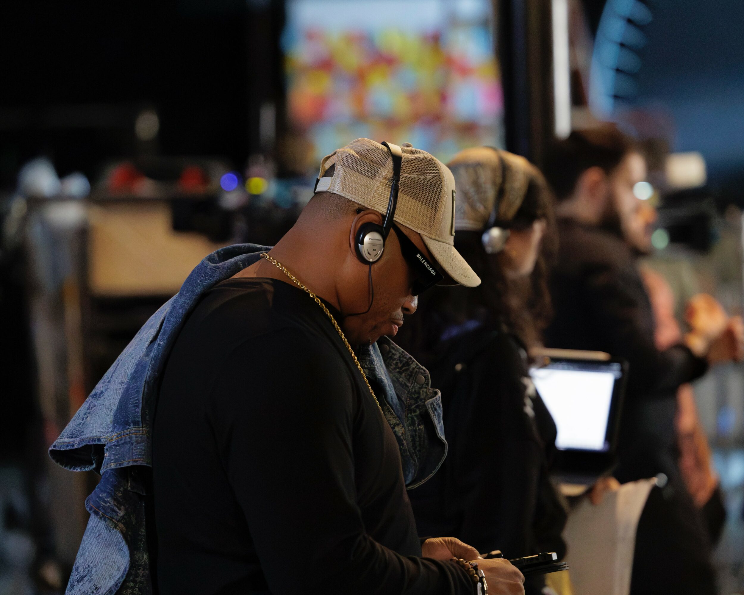 Person wearing a beige cap and headphones, looking down, in a busy indoor setting with others in the background.