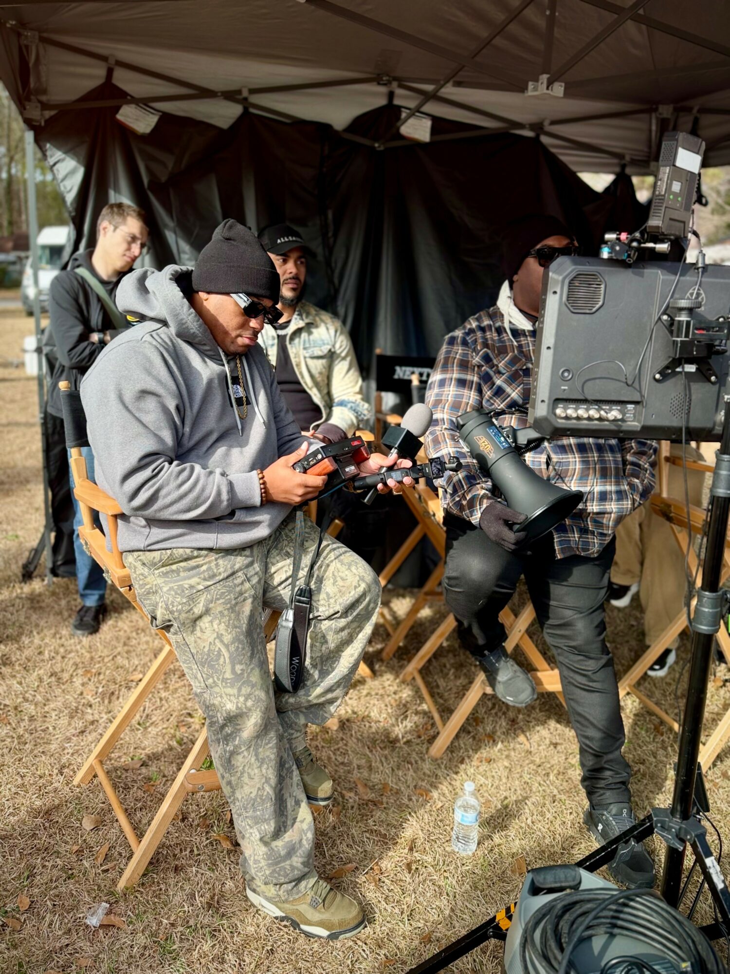 Group of people operating camera equipment outdoors under a tent, with a black backdrop and a water bottle on the ground.