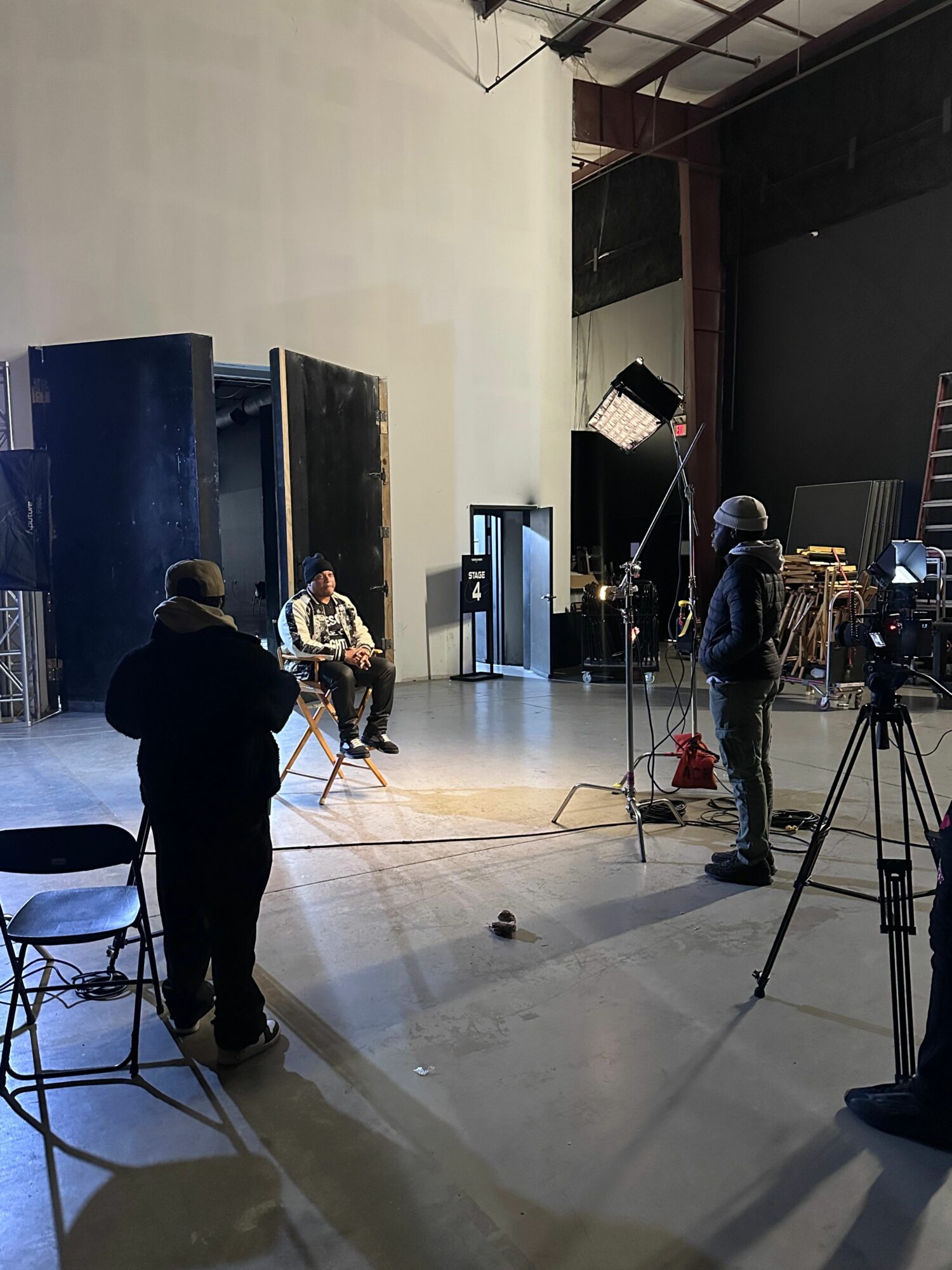 People in a studio with equipment, one person sitting on a chair, another standing, and a person working with a camera.