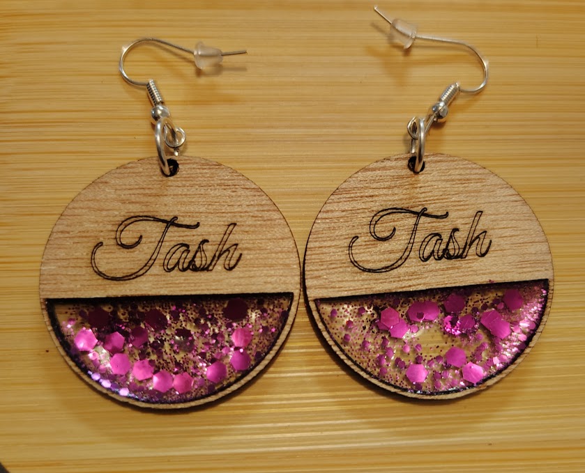Round wooden earrings with 'Task' written on top and pink glitter inside, on a wooden surface.