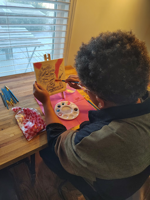 Child with curly hair and glasses paints at a table with art supplies, near a window with blinds.